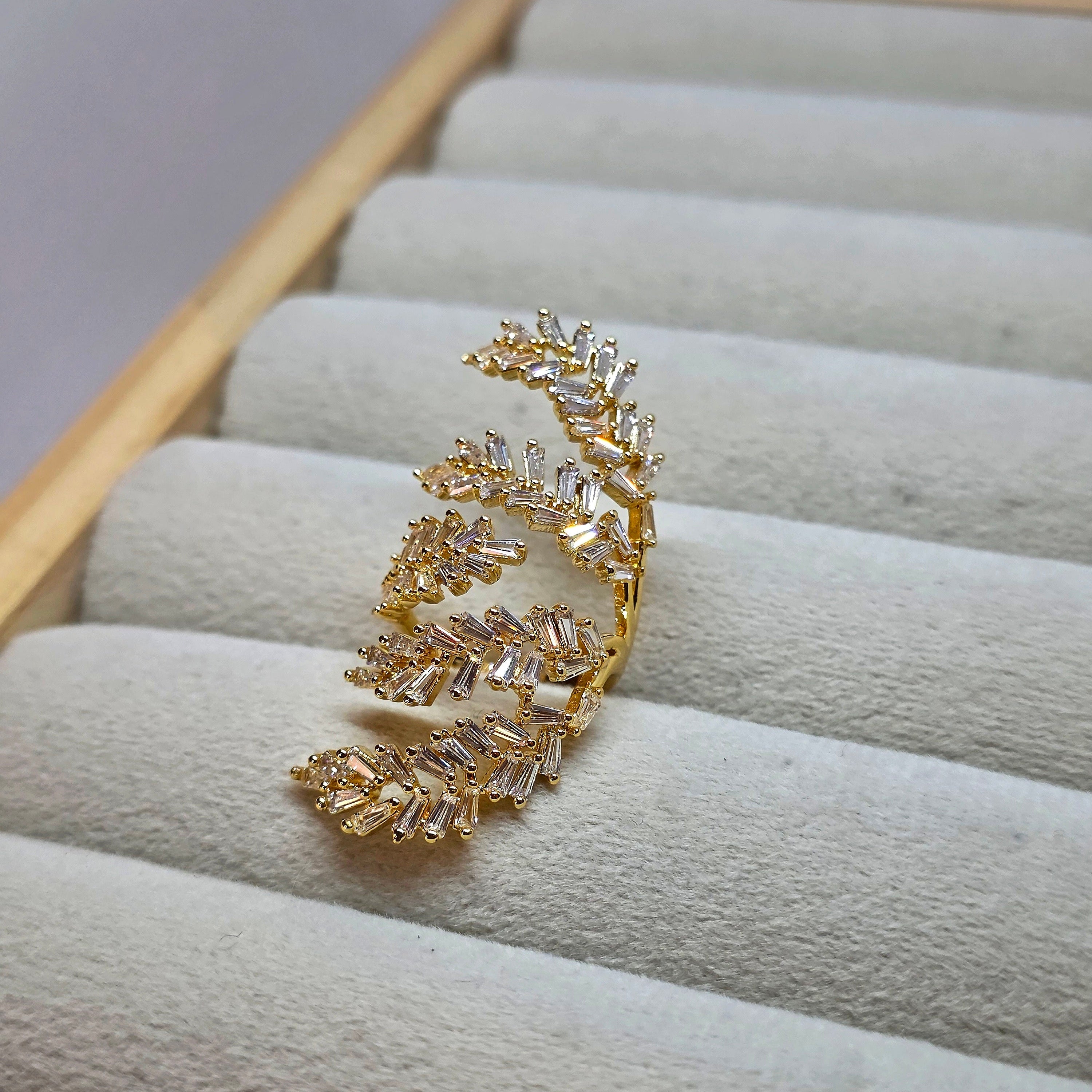 A gold leaf-shaped ring with clear baguette crystals, displayed on a light beige velvet jewelry organizer tray.
