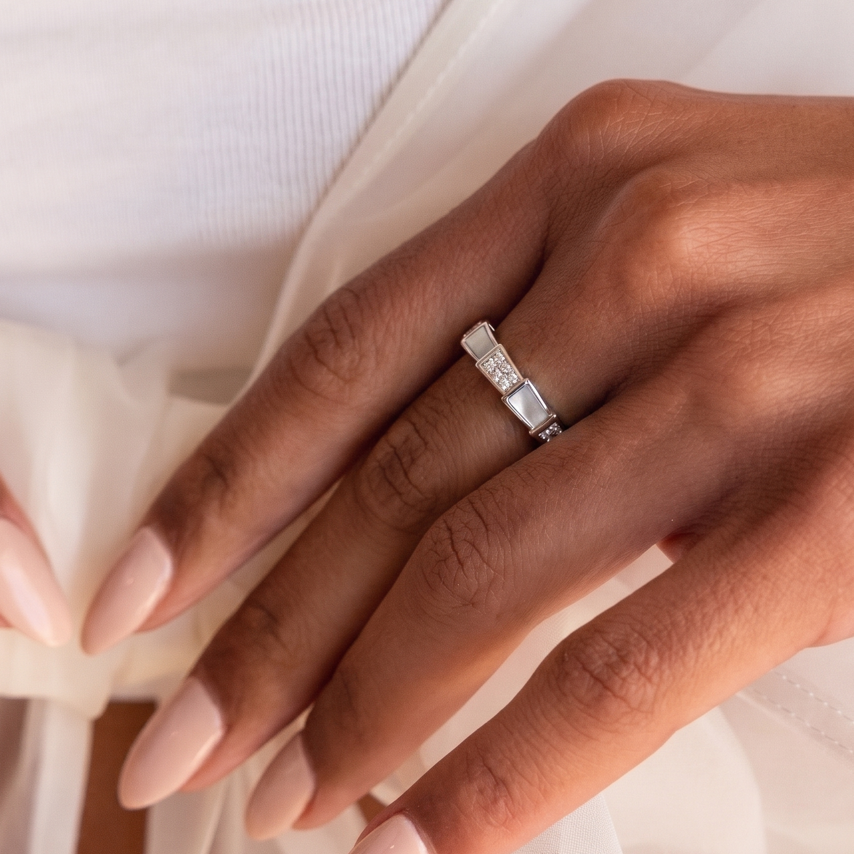 A close-up of a hand wearing a silver band ring with a row of small clear stones, styled with a white textured garment and sheer fabric.