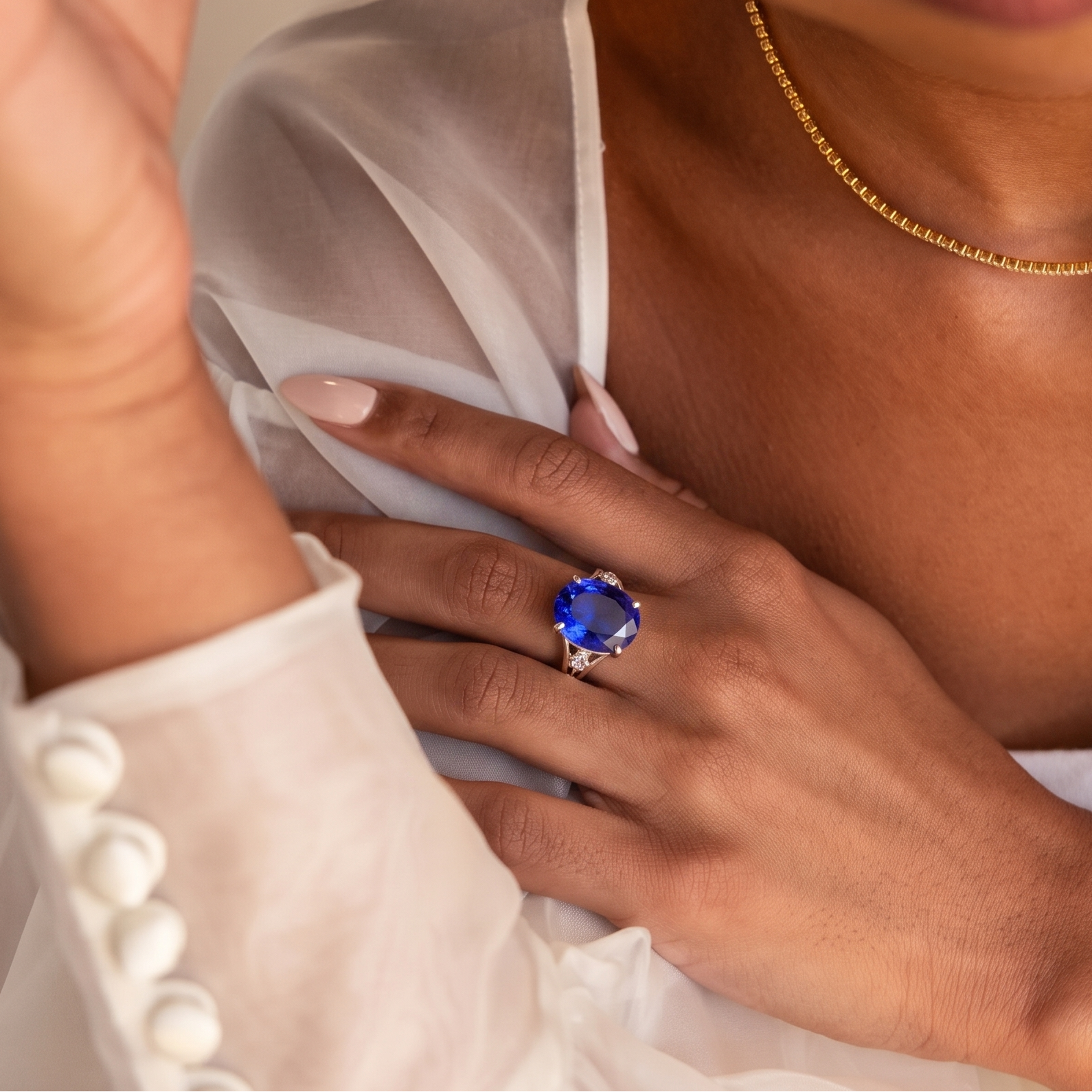 A close-up lifestyle shot of a model wearing a large oval-cut blue gemstone ring on her finger, styled with a delicate gold chain necklace and a white sheer blouse.