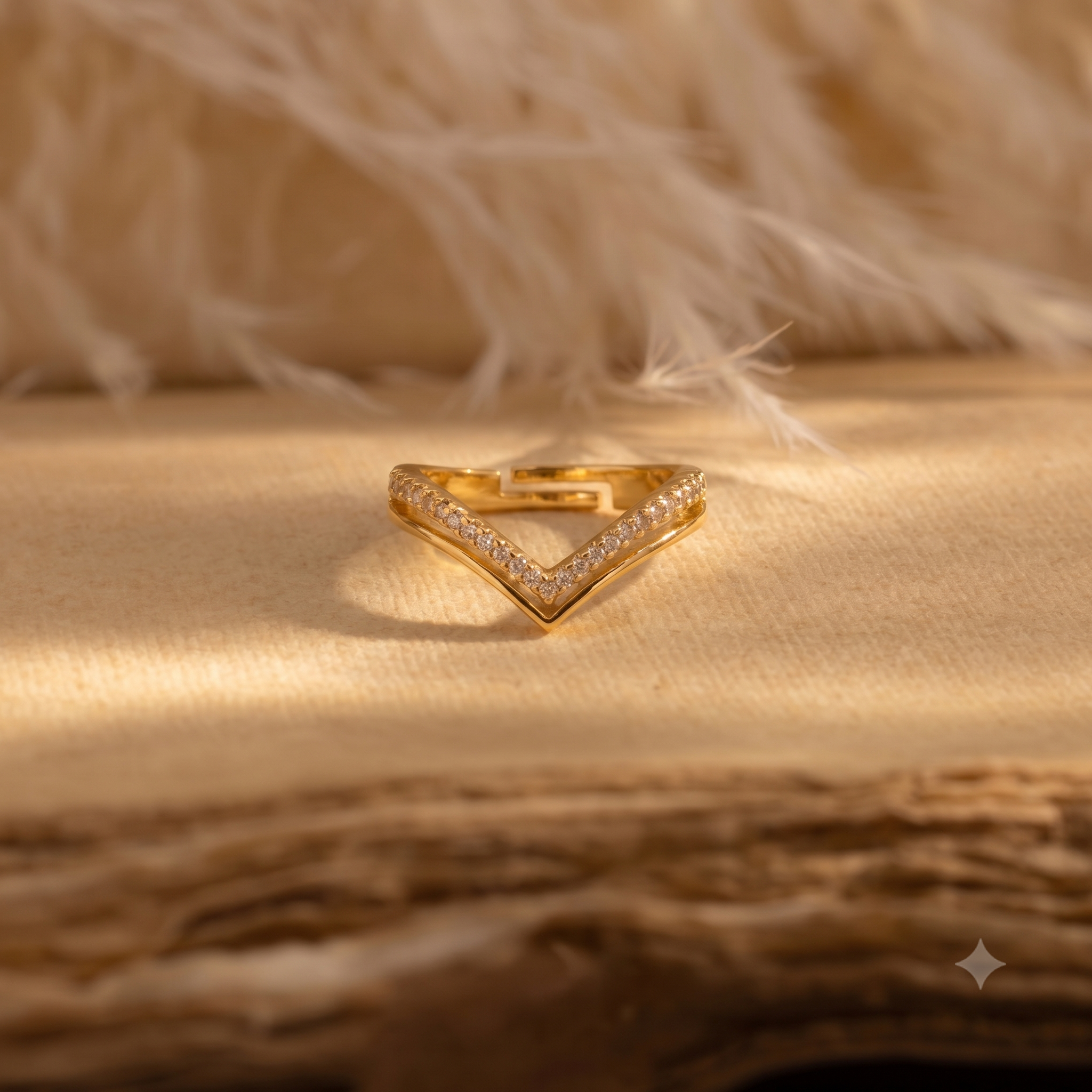 A gold V-shaped ring with pavé-set clear stones, resting on a textured beige surface with a soft, blurred feather background.