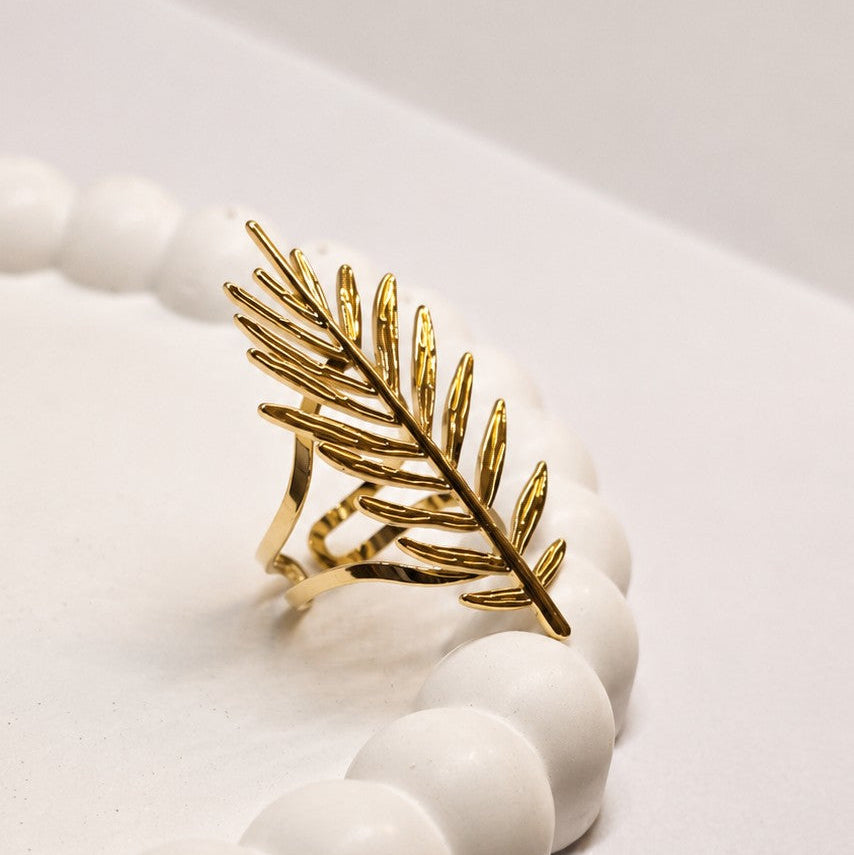 A gold leaf-shaped ring displayed on a white beaded surface against a clean white background.