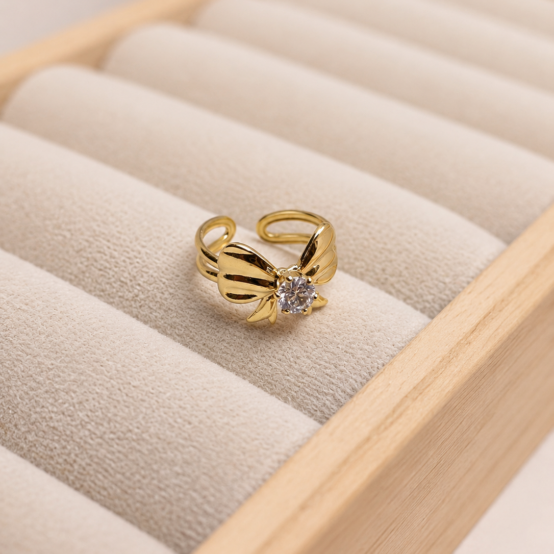 A gold ring with a stylized bow design and a clear central gemstone, displayed on a beige velvet jewelry box tray.