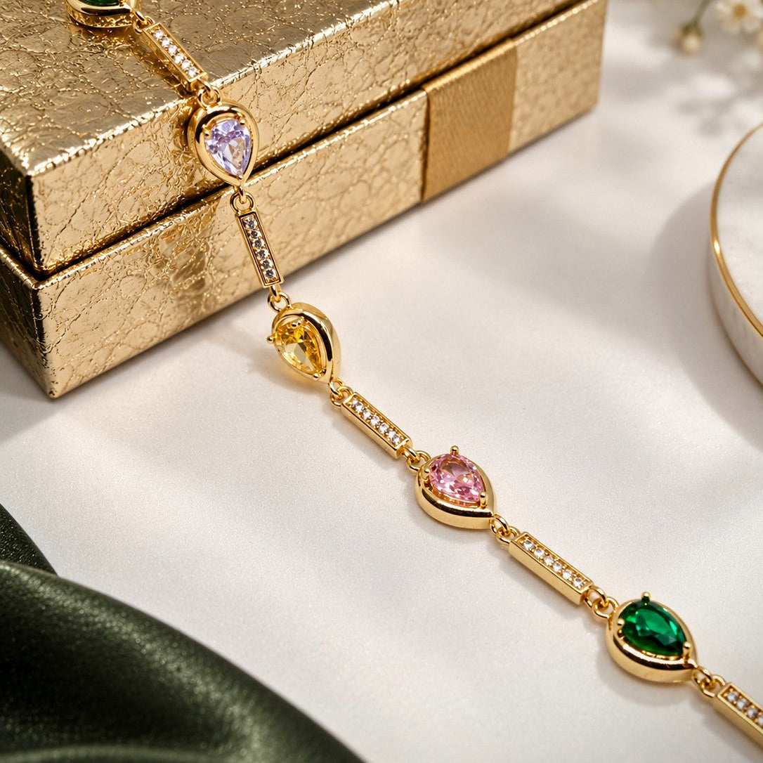 A gold-toned bracelet with multiple teardrop-shaped gemstone settings in green, purple, yellow, and pink, styled on a white surface next to a textured gold gift box.