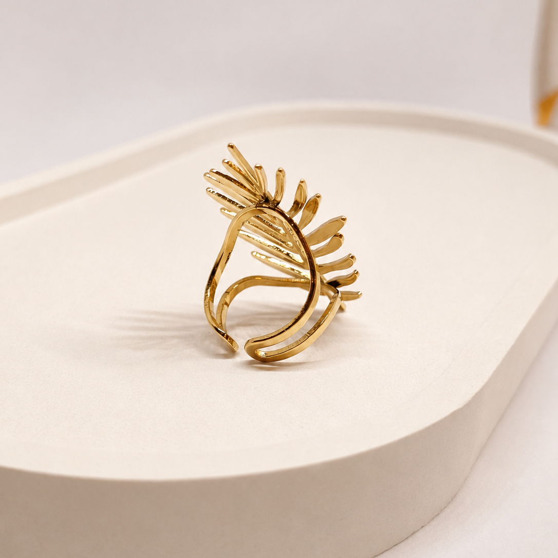 A gold-toned abstract ring with a sculptural, leaf-like or flame-like openwork design, displayed on a white curved surface.