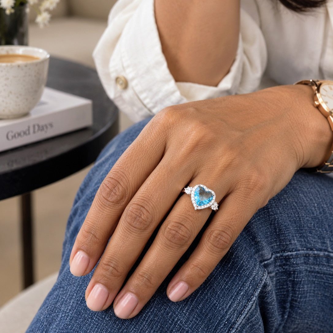 A close-up lifestyle shot of a person's hand wearing a silver heart-shaped ring with a light blue gemstone, styled with blue jeans and a white shirt in a cozy indoor setting.
