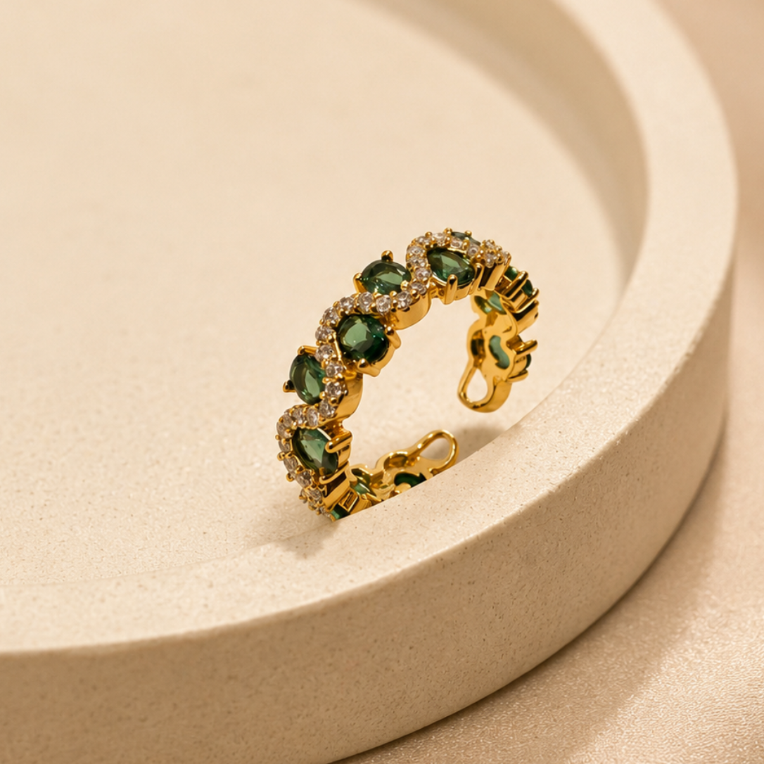 A gold ring with green gemstone accents, displayed on a beige textured circular pedestal.