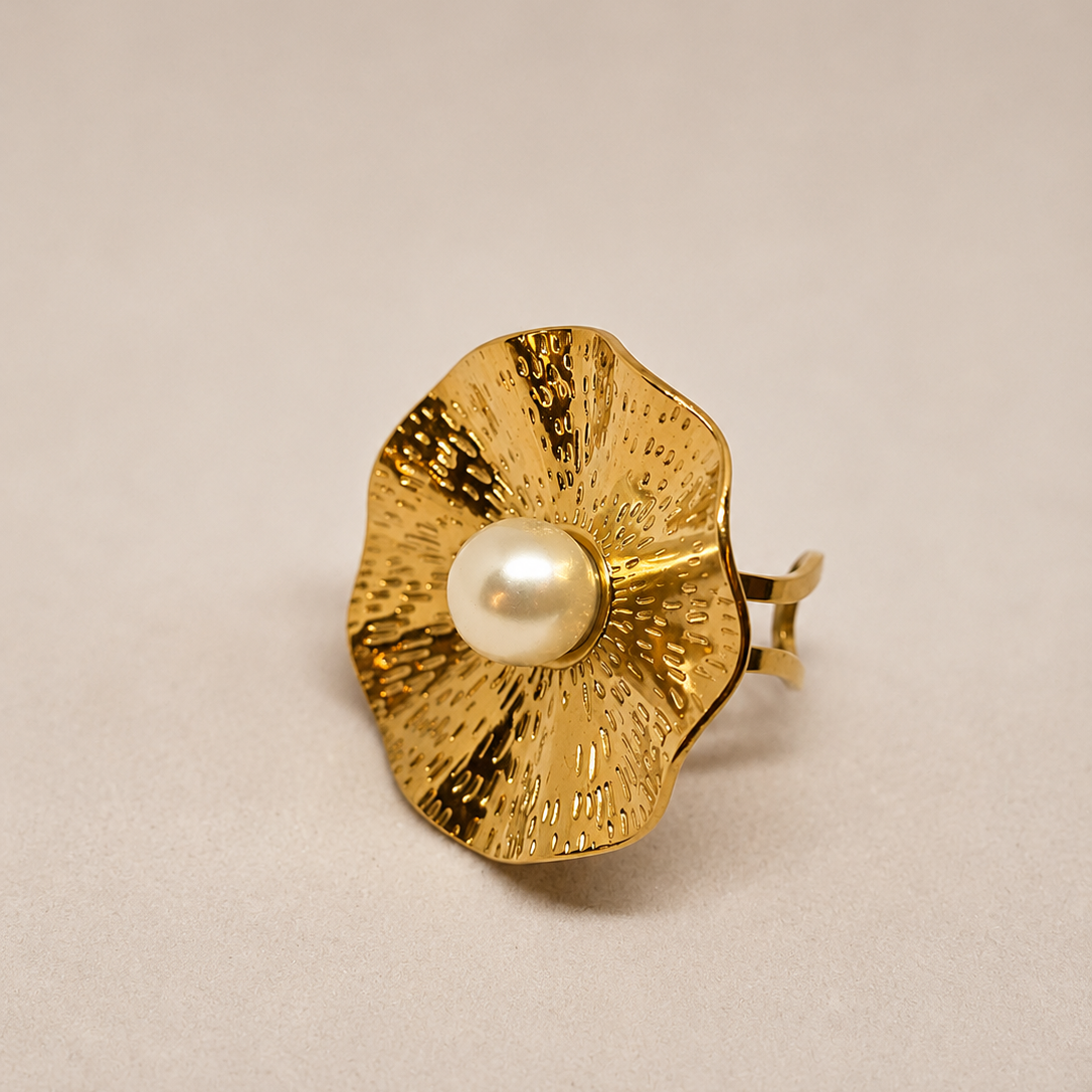A gold ring designed like a textured flower with a central white pearl, photographed on a neutral beige background.