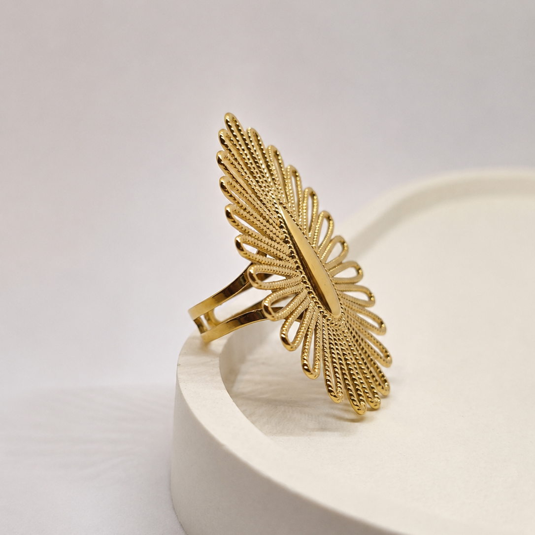 A gold-toned statement ring with a textured, fan-like leaf or sunburst design, displayed on a white ceramic pedestal against a light gray background.
