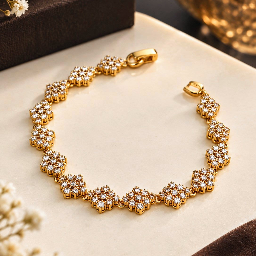 A gold bracelet with repeating floral clusters set with clear stones, displayed on a cream surface with decorative flowers and a dark box in the background.