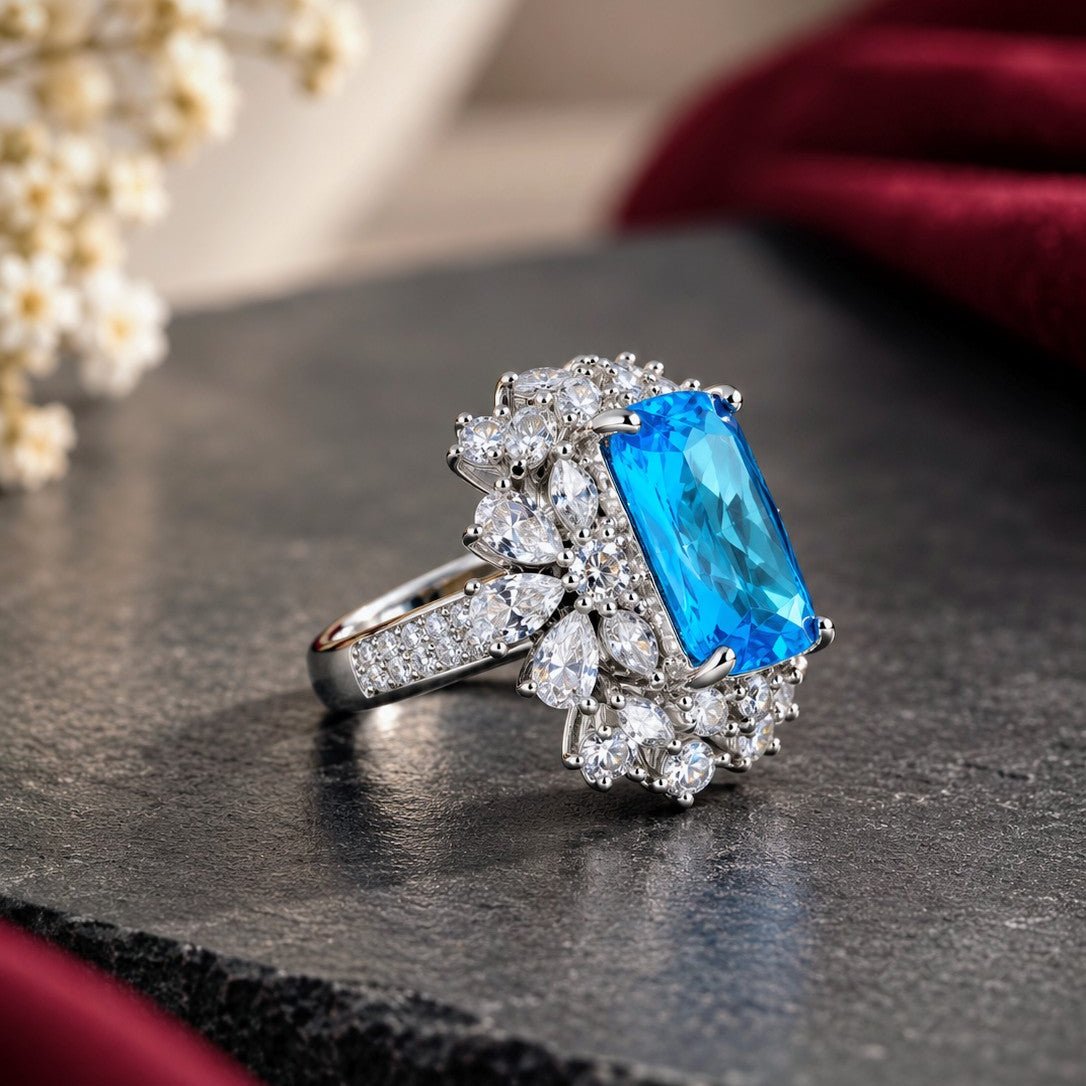 A silver-toned ring featuring a large rectangular blue gemstone surrounded by clusters of small clear stones, displayed on a dark textured surface with soft red fabric and dried flowers in the background.