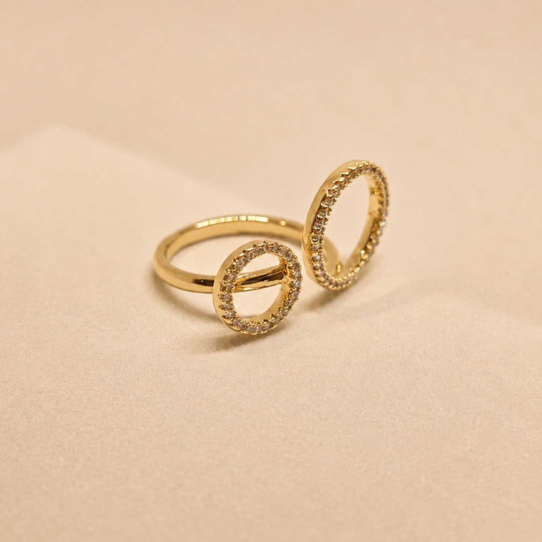A gold ring with two interlocking open circular designs, accented with small pavé-set clear stones, photographed on a warm beige background.
