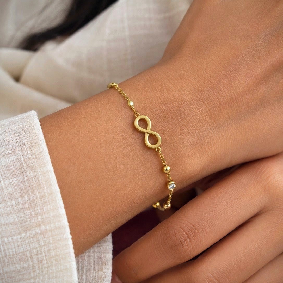 A delicate gold infinity bracelet with small bead accents and clear crystal details, shown worn on a person's wrist.