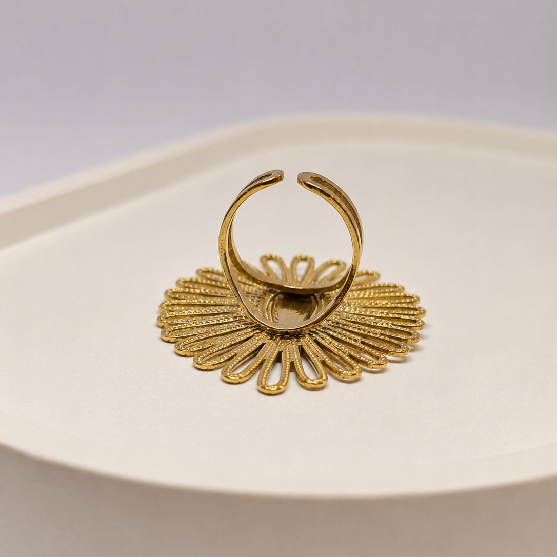A gold-toned metal ring with a textured, fan-like petal design and a curved open top band, displayed on a cream-colored ceramic surface against a light gray background.