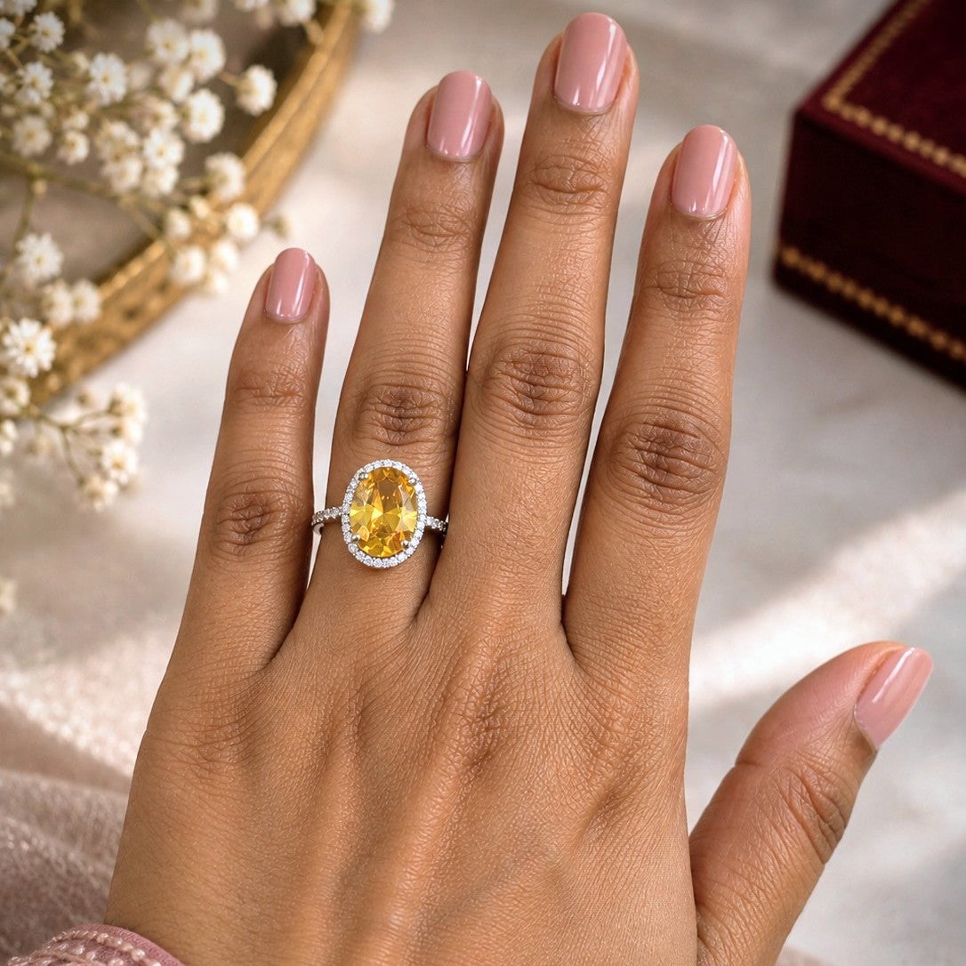 A close-up of a hand with pink manicured nails wearing an oval yellow gemstone ring with a silver halo setting, resting on a soft beige fabric background with dried flowers and a dark red box in the corners.