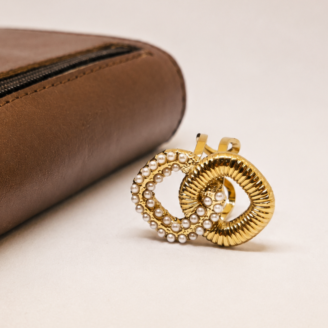 A gold-toned double-ring stud earring embellished with clear rhinestones, displayed next to a brown leather zippered pouch on a light beige background.