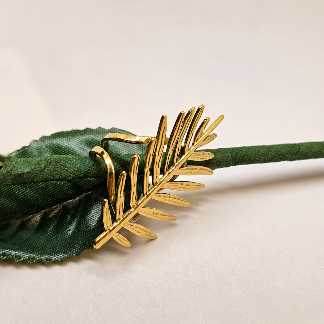 A close-up of a gold-toned leaf-shaped hair claw clip attached to a dark green fabric rose stem on a plain light background.