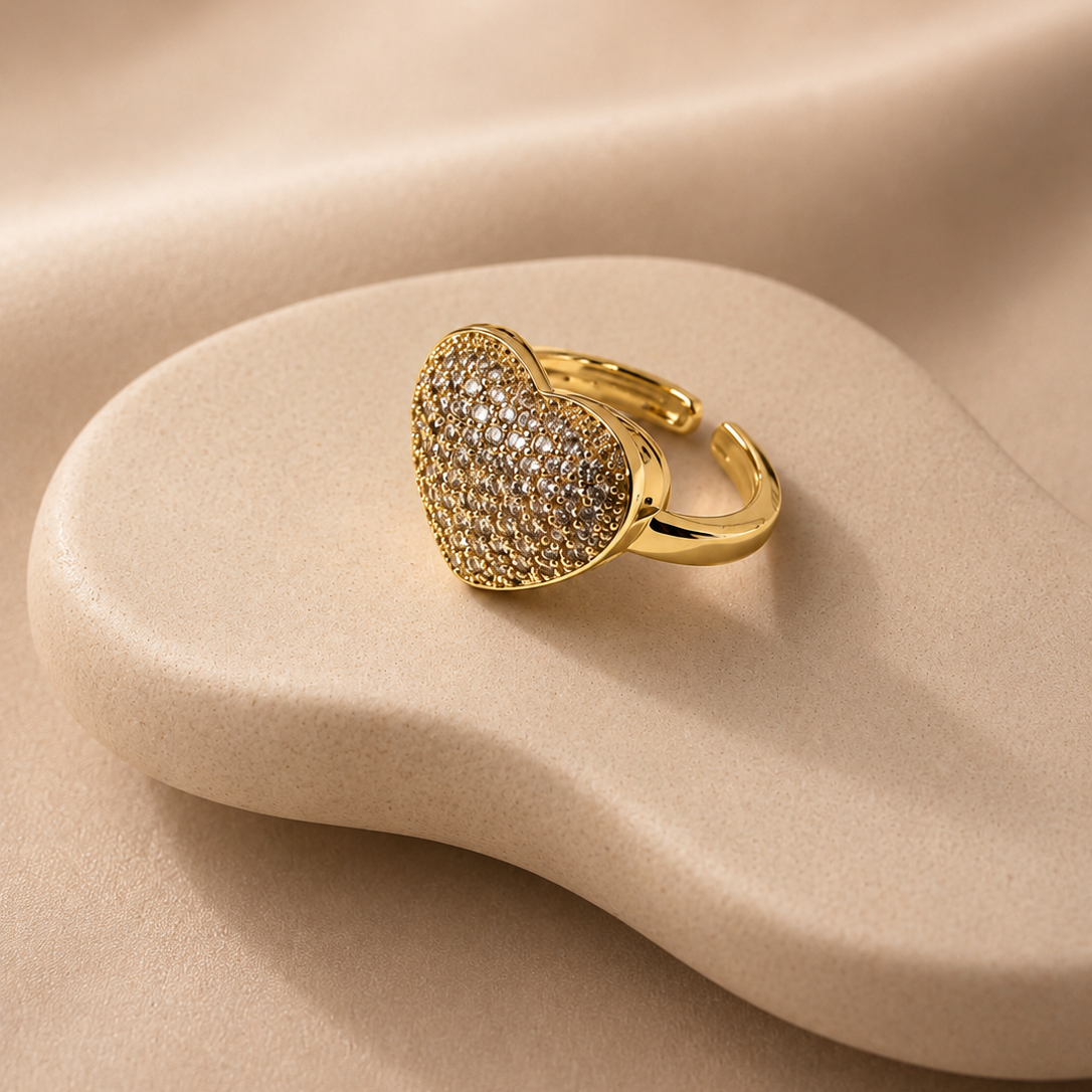 A gold heart-shaped ring with a textured pavé setting, displayed on a beige wavy stone prop against a draped beige fabric background.