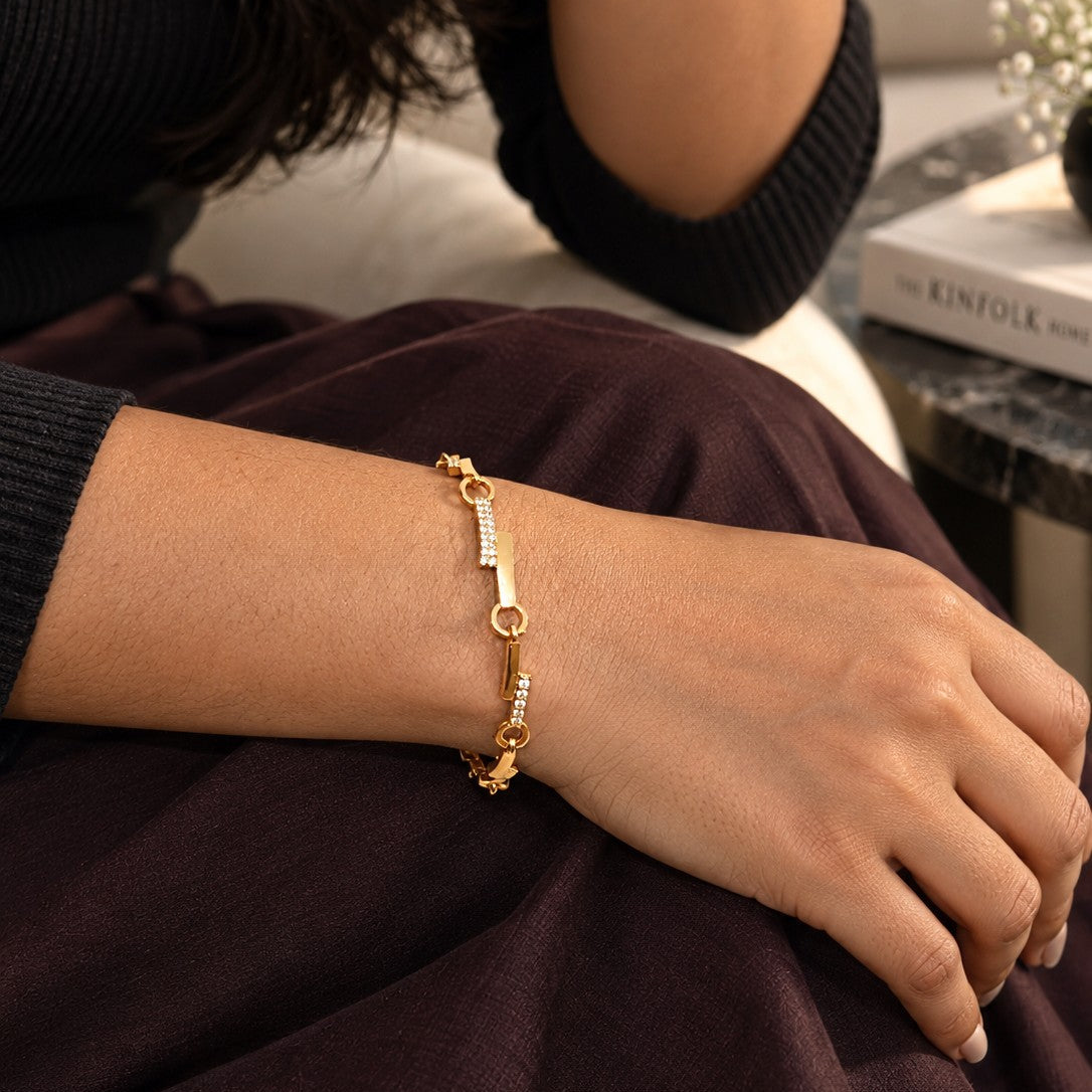 A gold chain bracelet with bar and bead details worn on a person's wrist, shown in a close-up lifestyle shot with a dark brown skirt and black ribbed top.