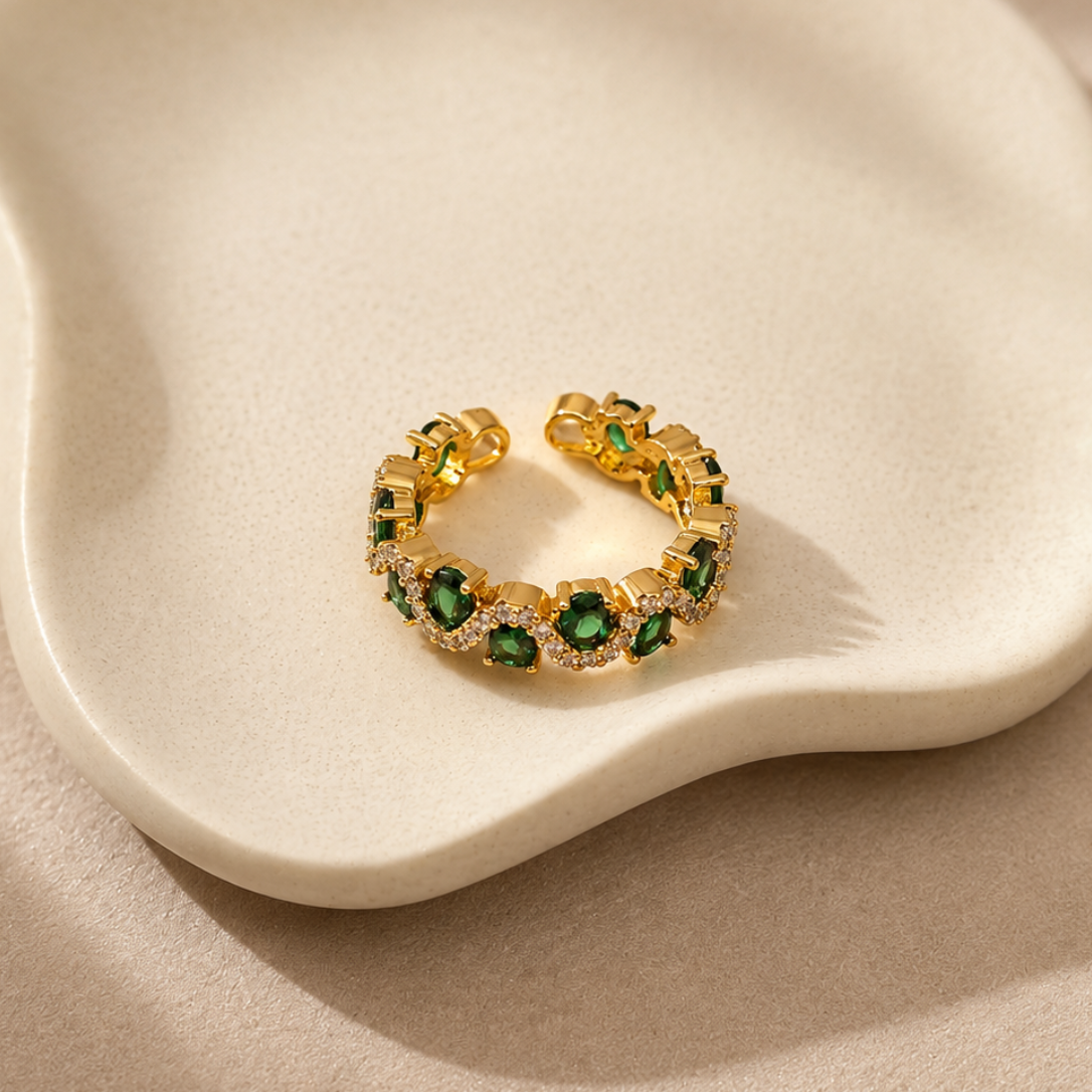 A gold ring with green gemstone clusters and small clear stones, displayed on a cream-colored ceramic dish against a beige fabric background.