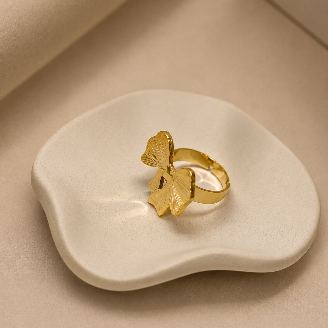 A gold ring with a textured leaf-shaped top, displayed on a small, organic-shaped cream ceramic dish against a warm beige background.