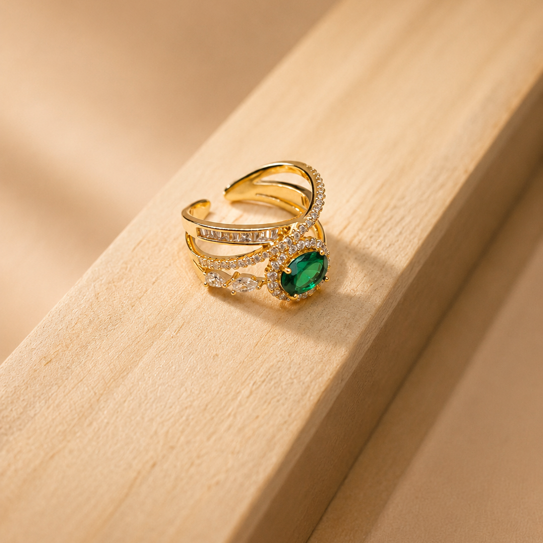A gold-plated adjustable ring with a green emerald-cut center stone and pavé-set clear stones, resting on a light wood surface.