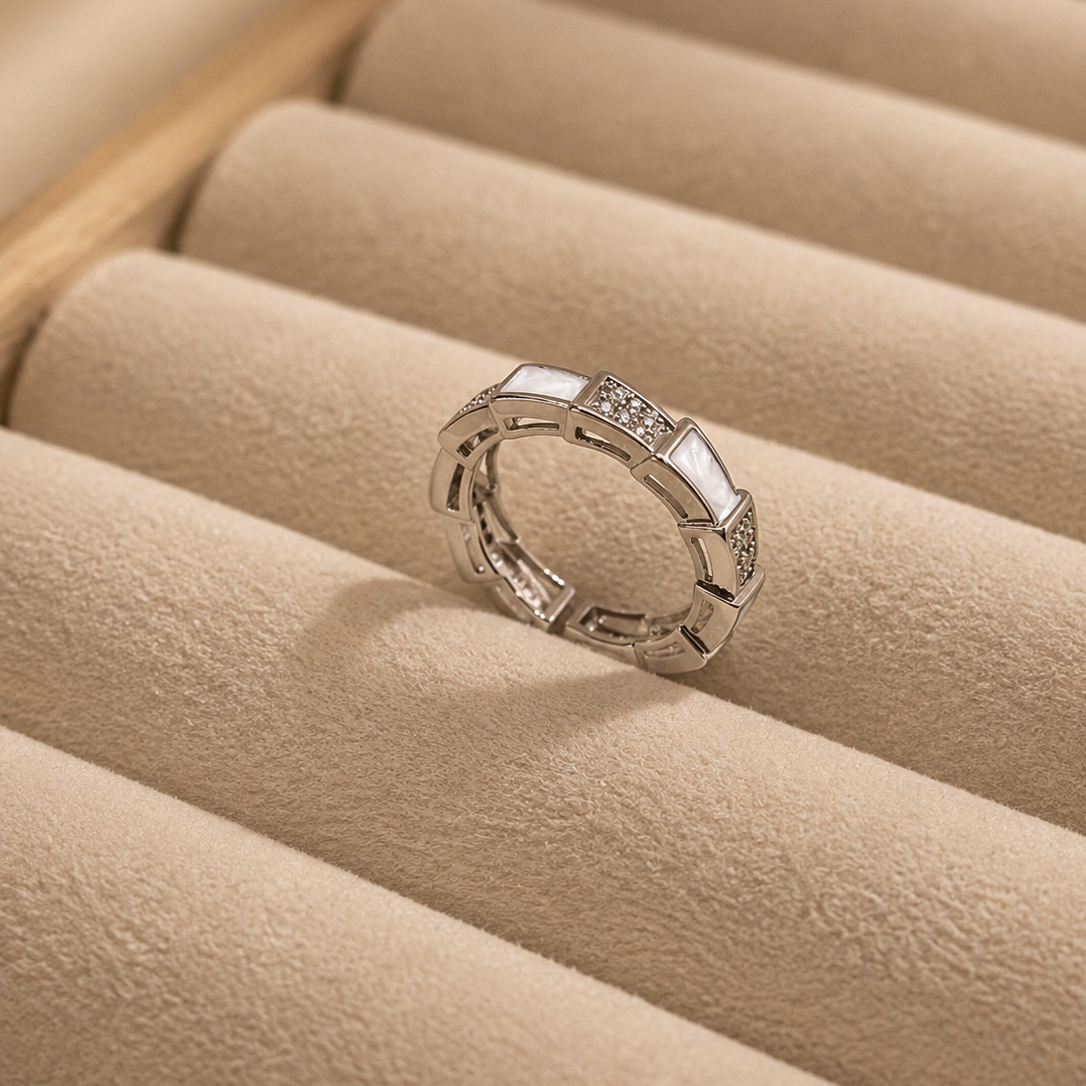 A silver-toned geometric ring with a small clear gemstone, displayed on a beige textured jewelry tray.