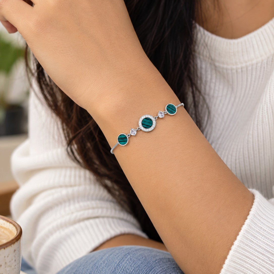 A close-up of a woman's wrist wearing a delicate silver bracelet with three green oval stones and a small clear center stone, styled with a white ribbed sweater and blue jeans.