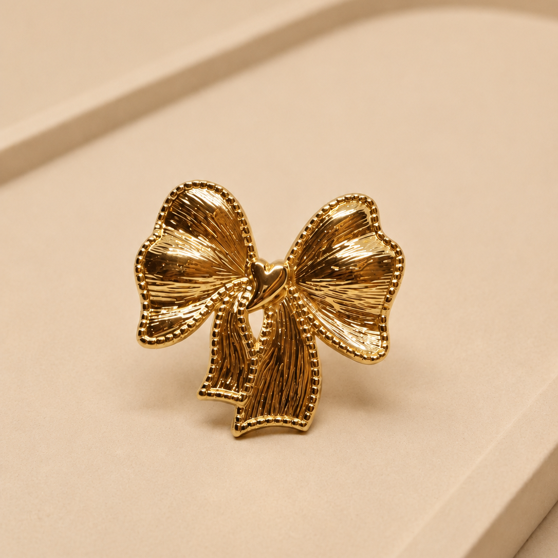 A gold bow-shaped ring with a textured, ribbed surface and beaded edges, displayed on a beige surface.