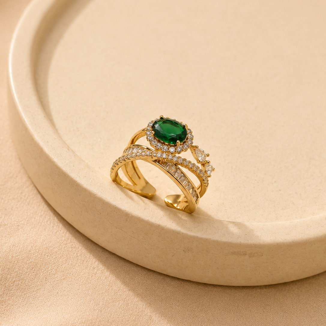A gold ring with a round green emerald-cut center stone and pavé-set clear accent stones, displayed on a beige ceramic tray against a beige fabric background.