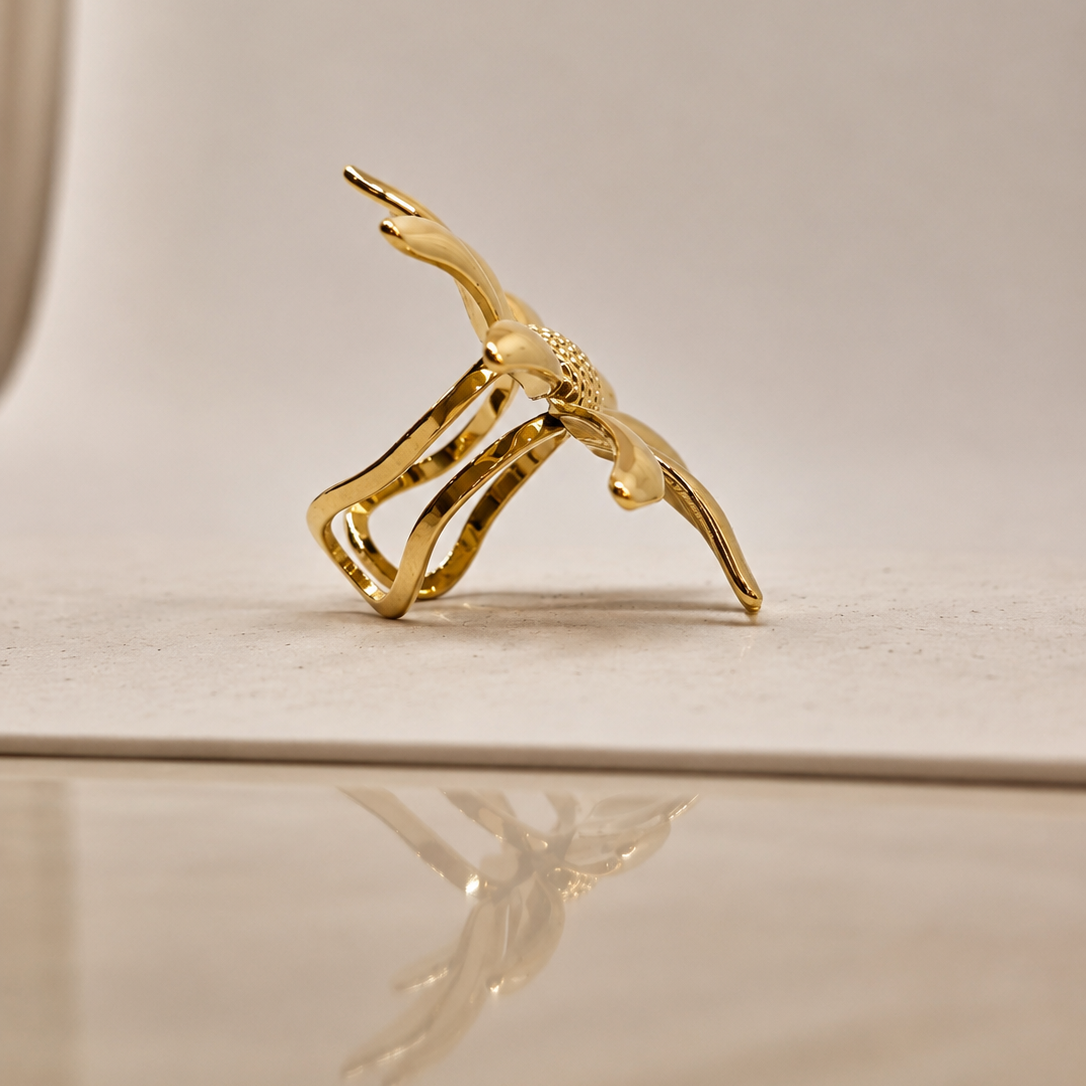 A small gold-toned sculptural ring or jewelry piece shaped like a stylized human figure, photographed on a warm beige surface with a soft reflection below.