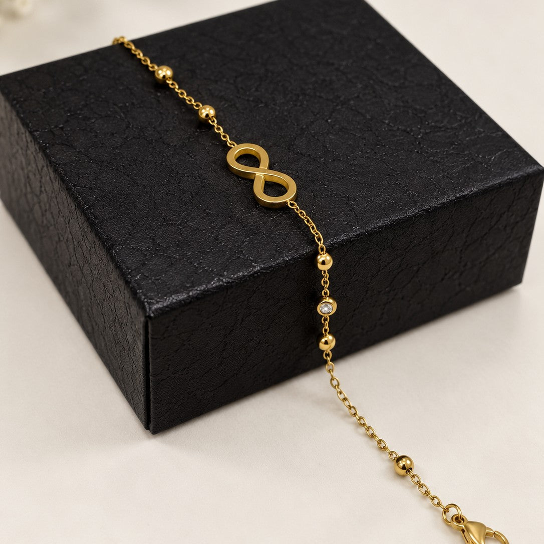 A delicate gold chain bracelet featuring a central infinity symbol and small bead accents, draped over a textured black gift box on a light beige surface.