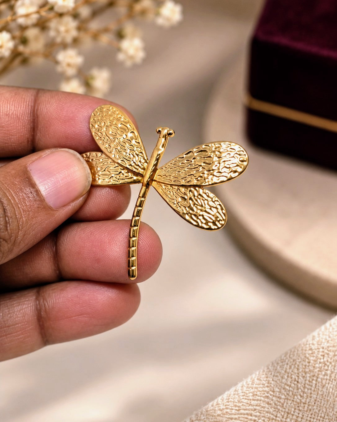 A hand holding a gold dragonfly brooch with textured wings, set against a soft beige background with a blurred velvet box in the corner.