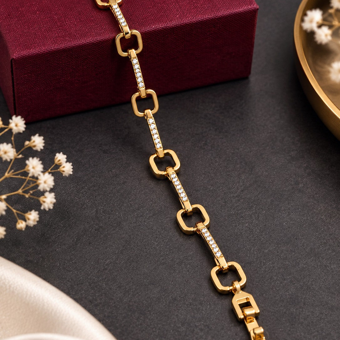 A gold-tone chain bracelet with alternating open-link and pavé-set stone links, displayed diagonally on a dark textured surface next to a burgundy jewelry box and small white flowers.