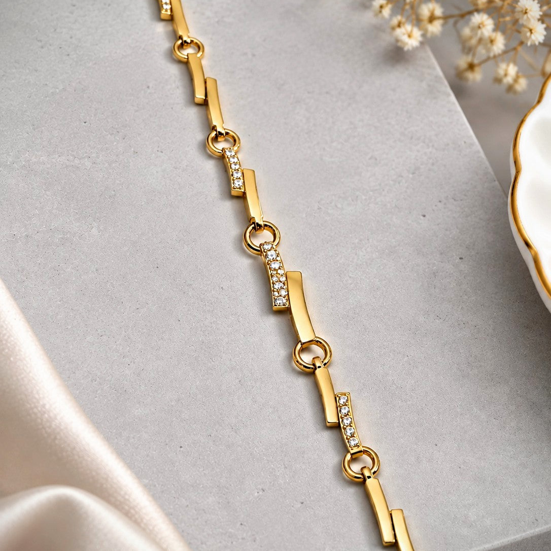 A gold link bracelet with geometric bar and ring motifs, some embellished with small clear stones, displayed diagonally on a gray surface with satin fabric and dried flowers as props.