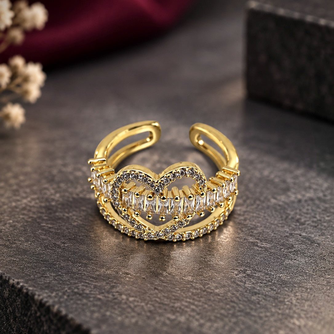 A gold adjustable ring with a heart-shaped design set with clear crystals, displayed on a dark textured surface with a blurred dark red fabric and small dried flowers in the background.