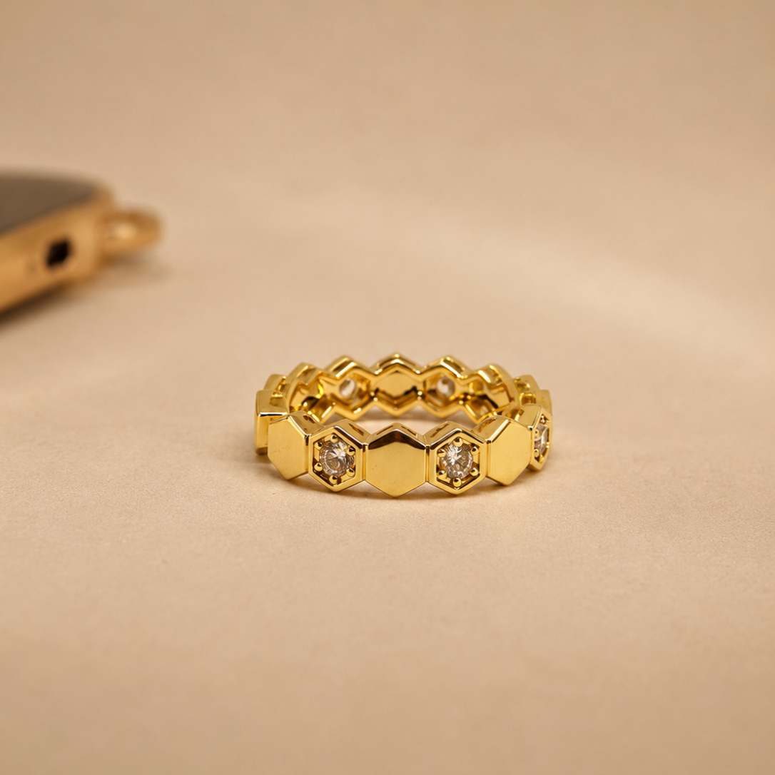 A gold honeycomb ring with small clear stones set in the hexagonal gaps, photographed on a warm beige background.