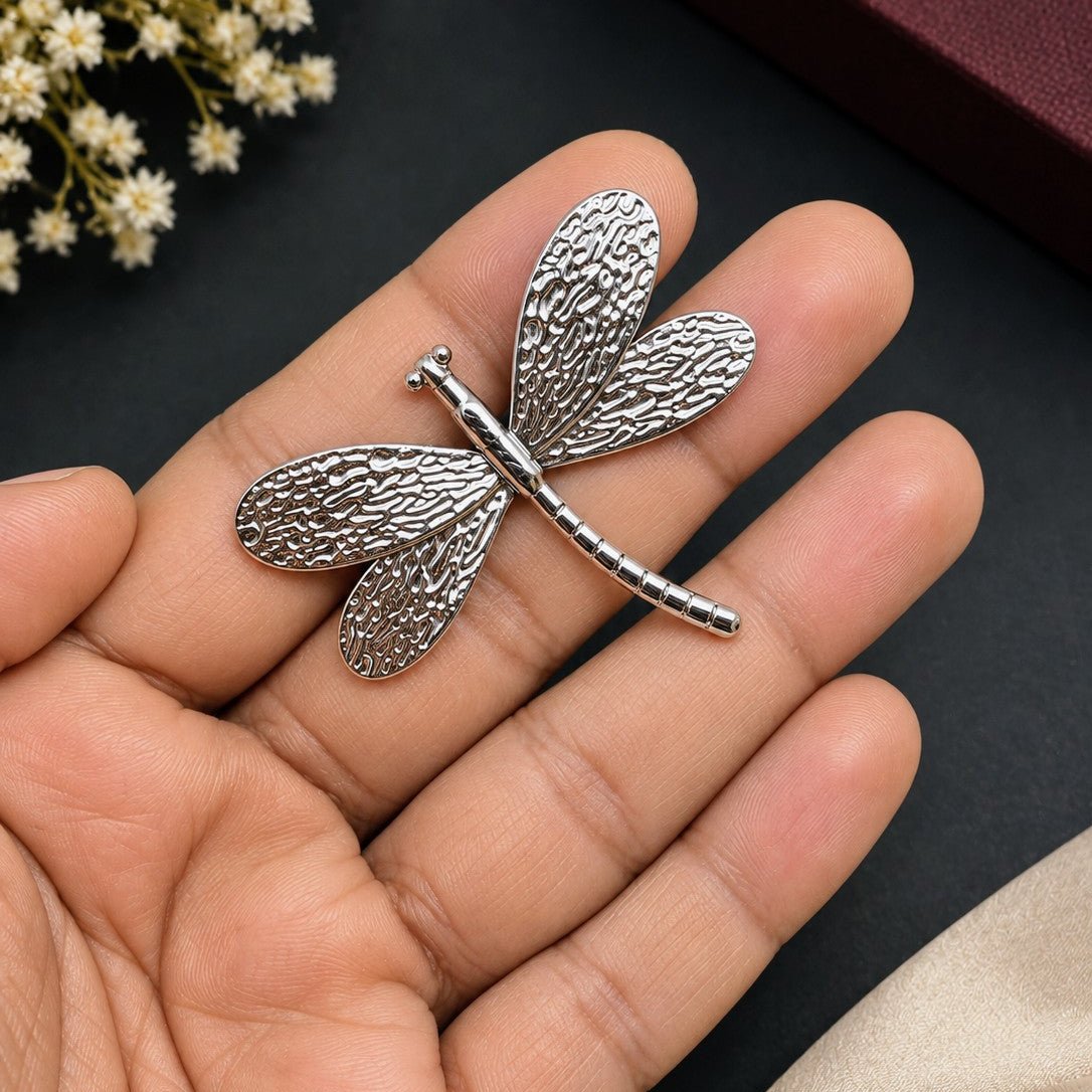 A silver dragonfly-shaped brooch with a textured, hammered finish resting on a person's hand, with a dark brown box and white flowers in the background.