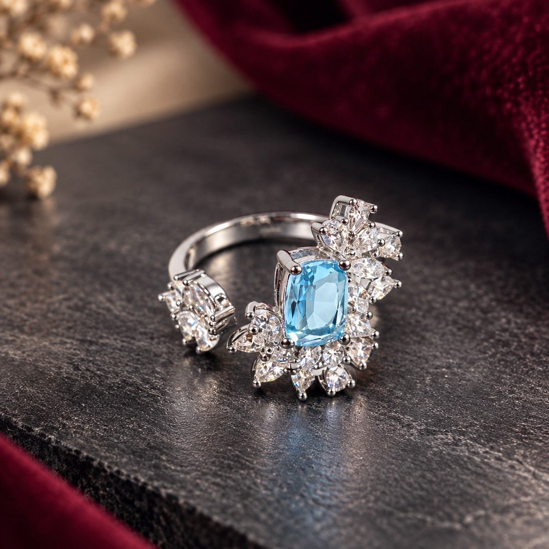 A silver-toned ring featuring a central light blue rectangular gemstone surrounded by clear stones in a floral cluster, displayed on a dark textured surface with red velvet fabric in the background.