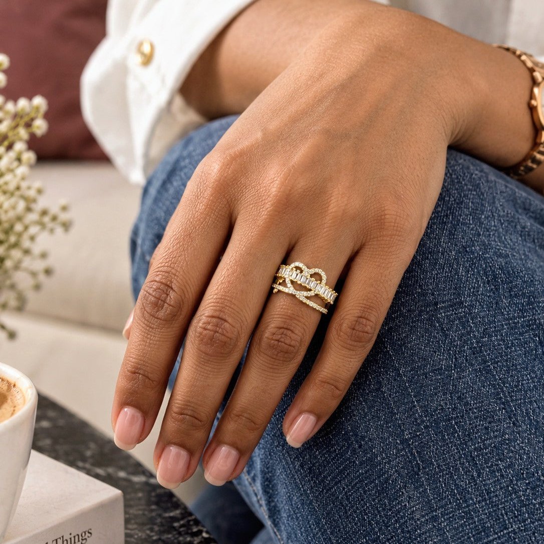 A close-up lifestyle shot of a person's hand resting on their lap, wearing a gold ring with a crown motif and small clear stones. The person is wearing a white button-down shirt and blue jeans, with a cozy indoor background.