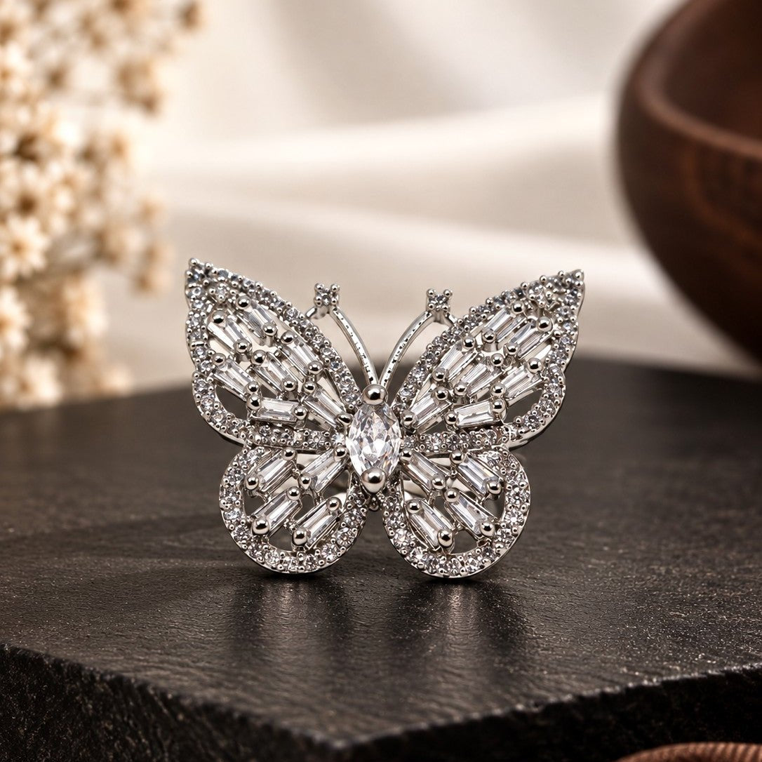 A butterfly-shaped silver ring set with clear marquise and round gemstones, displayed on a dark textured block with soft-focus decorative elements in the background.