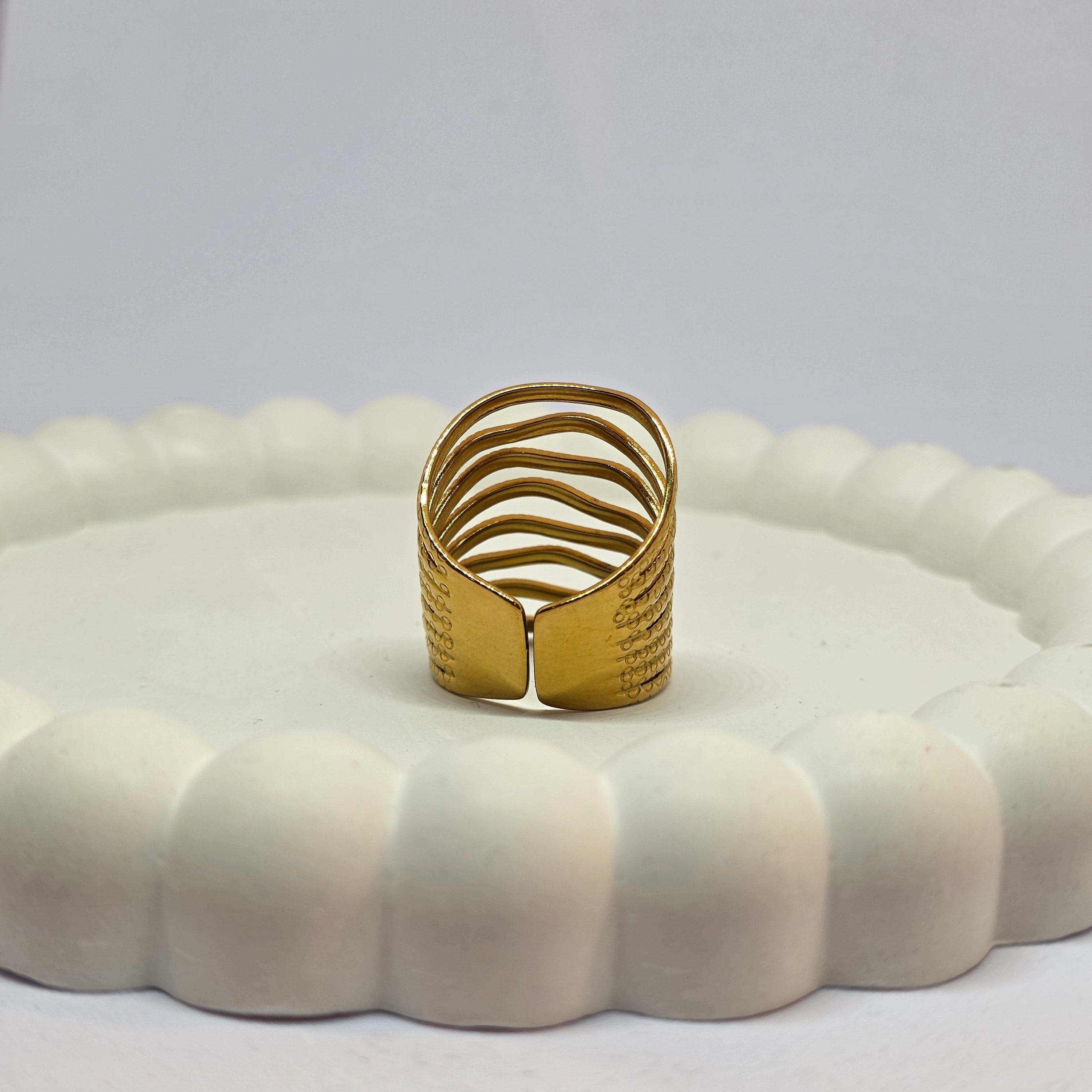 A gold-colored textured ring with a wavy, layered design, displayed on a white scalloped ring holder against a light gray background.