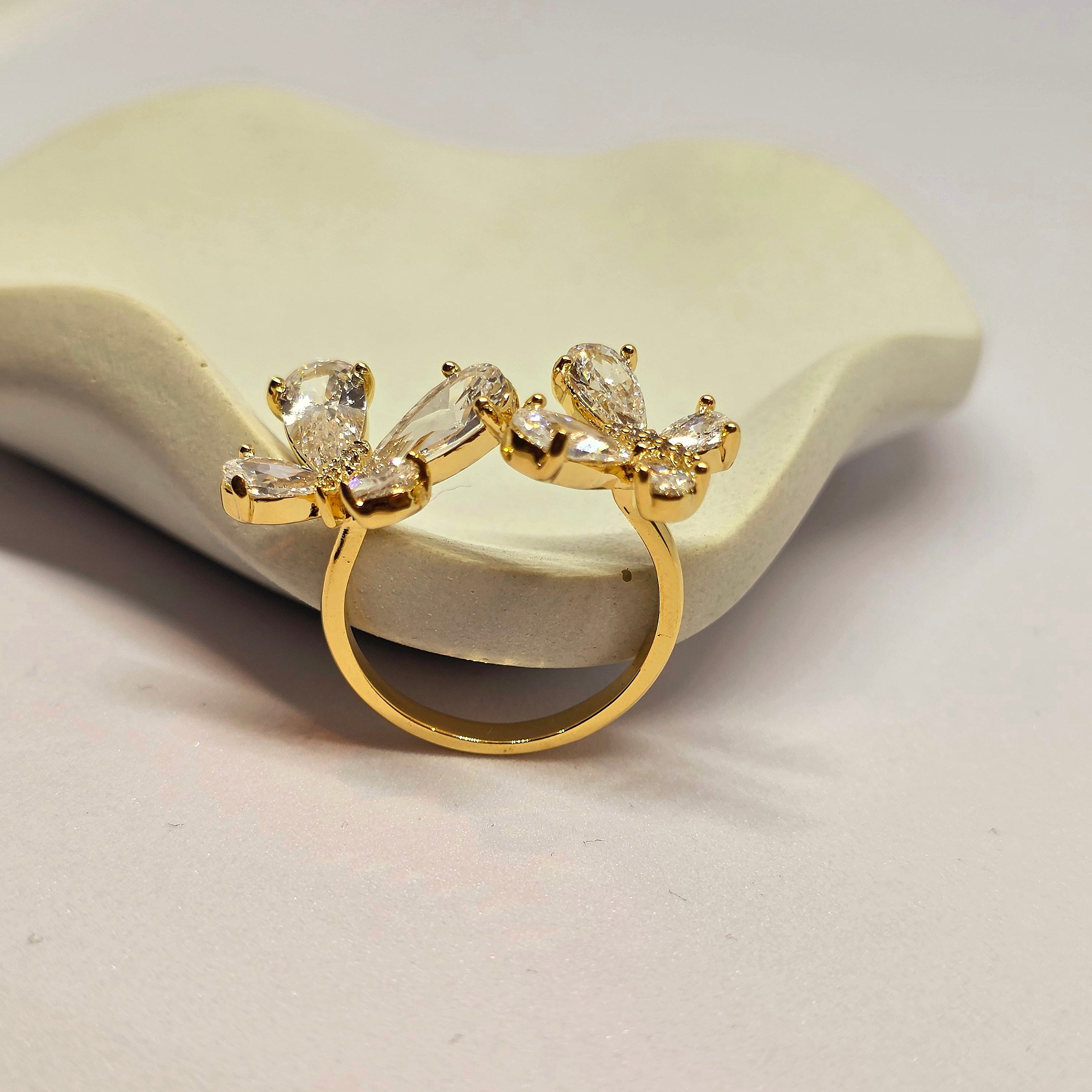 A gold ring featuring two butterfly-shaped clusters set with clear baguette and round gemstones, displayed on a light beige ceramic stand against a white background.