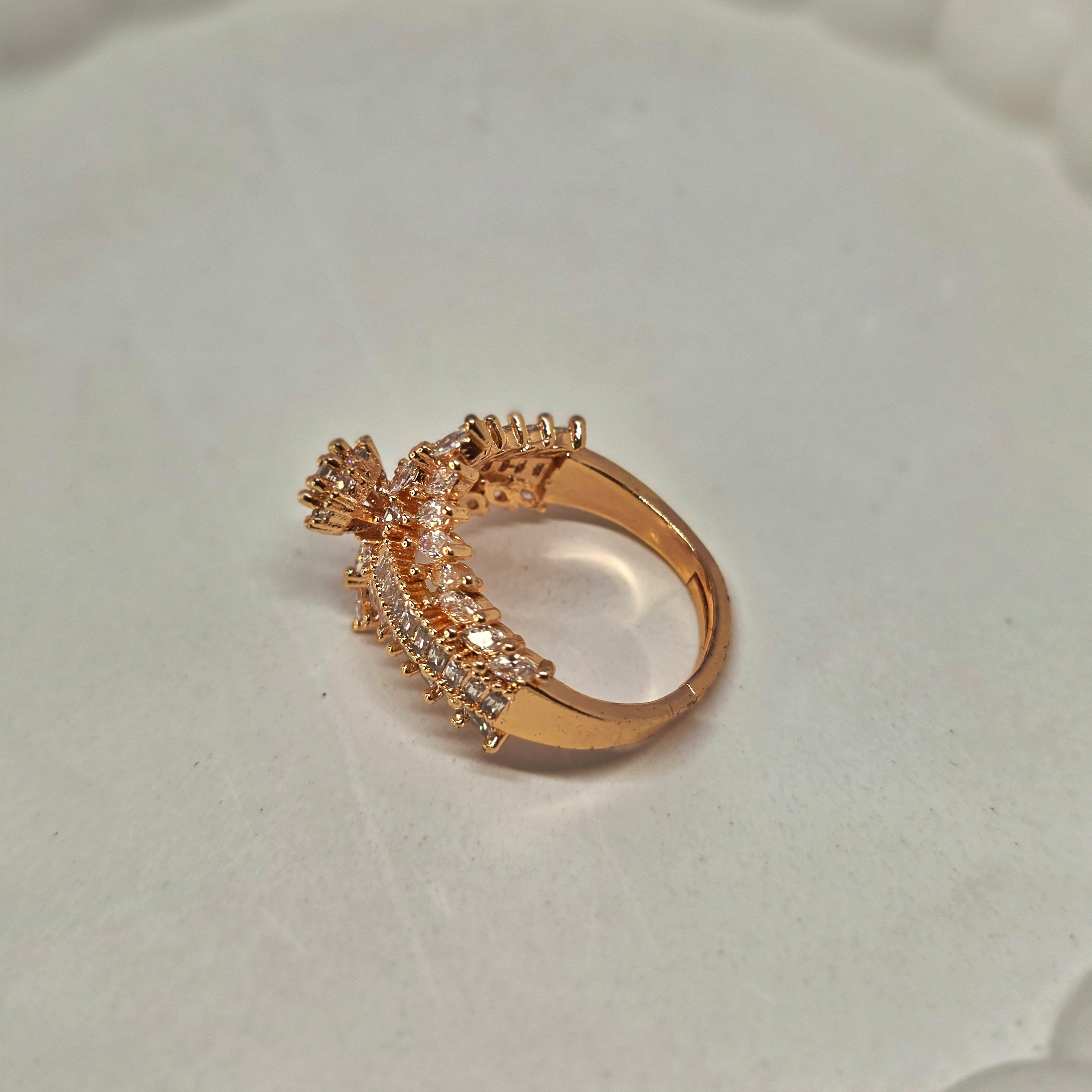 A rose gold ring with a floral cluster setting and pavé accents, resting on a white textured surface with a blurred beaded background.