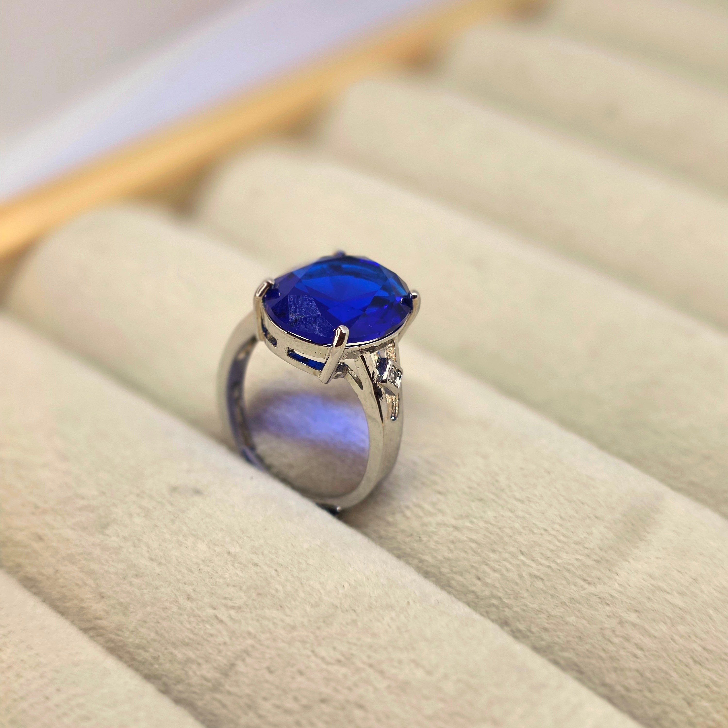 A silver ring with a large oval-cut deep blue gemstone, displayed on a beige velvet ring box.