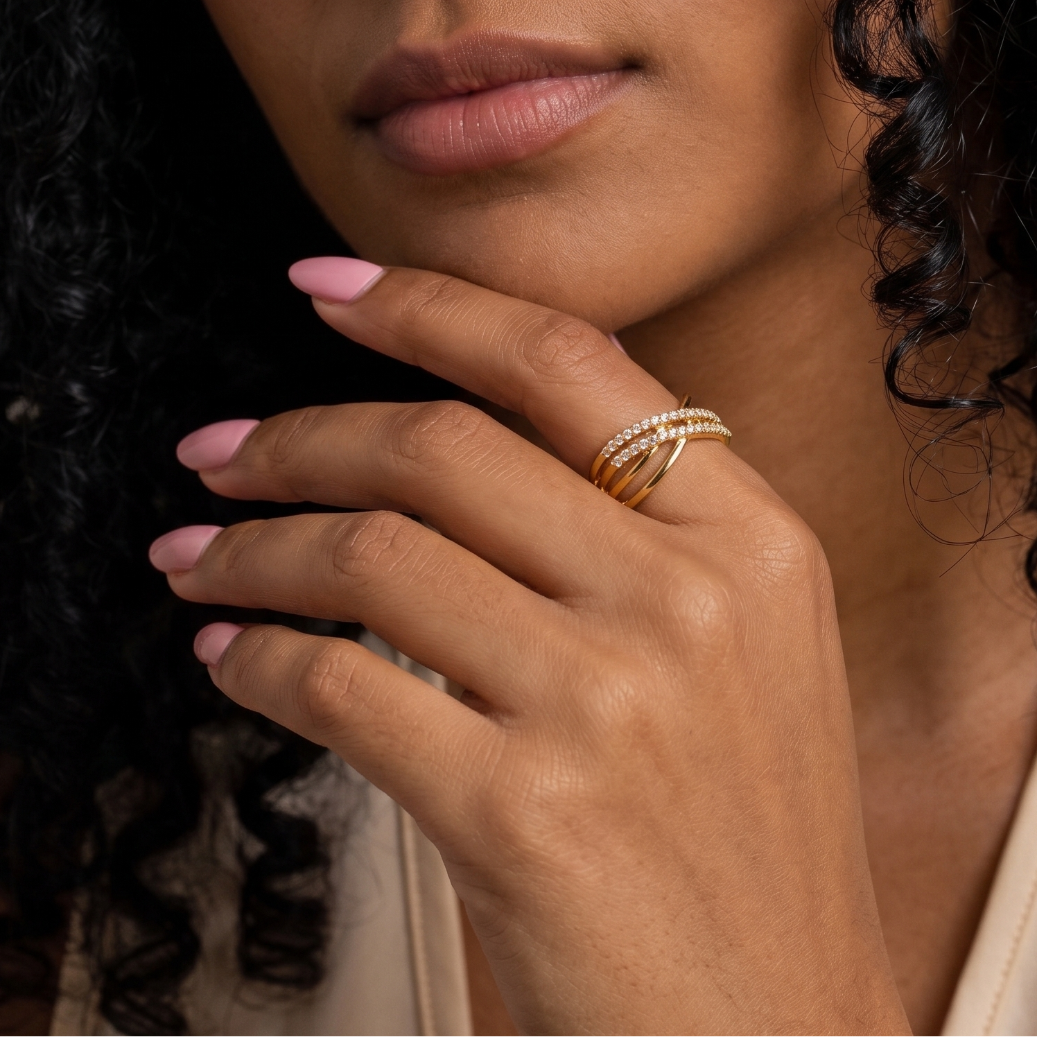 A close-up of a woman's hand wearing a delicate gold ring with small clear stones, set against her face and dark curly hair.