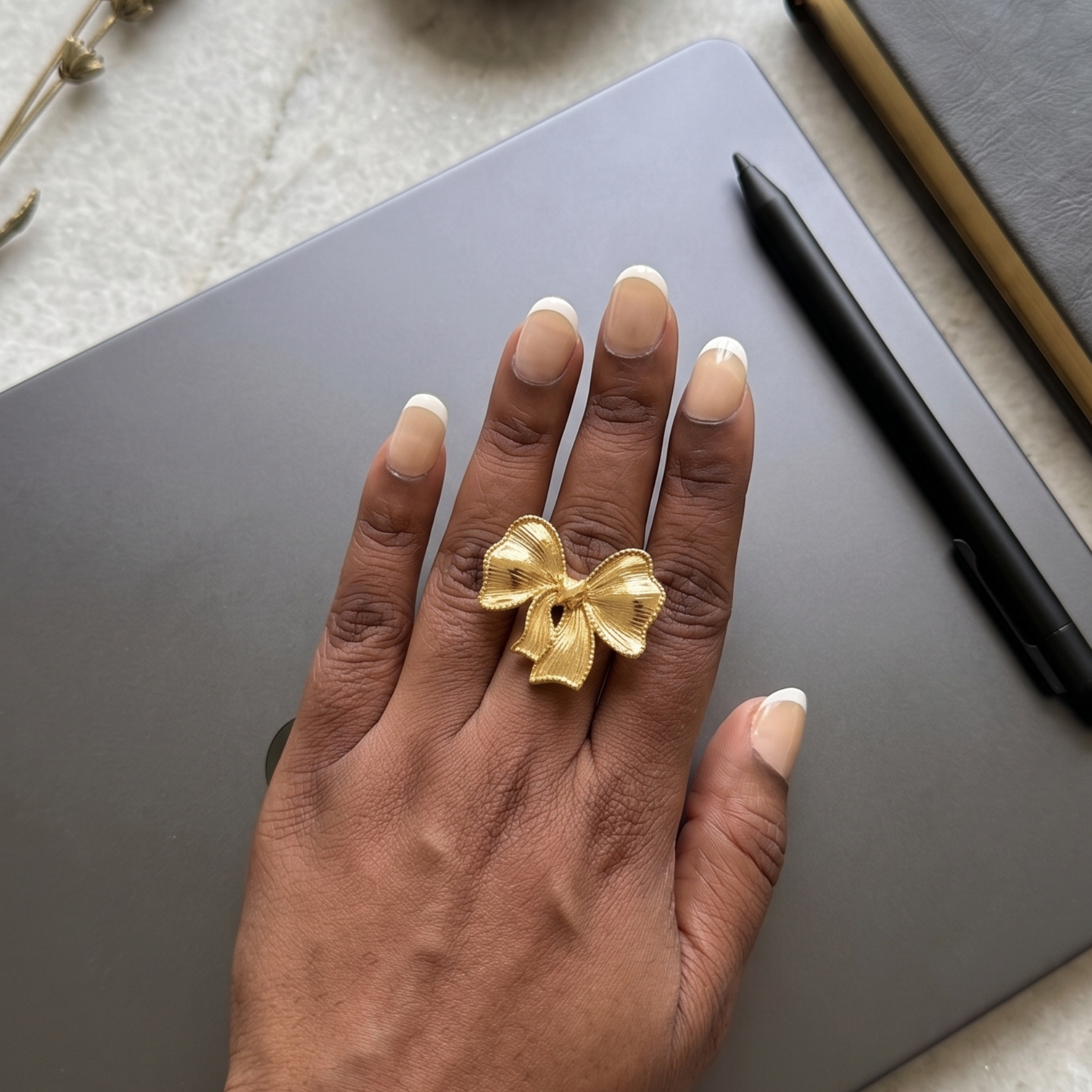 A gold ring shaped like a bow on a person's ring finger, shown resting on a dark gray tablet on a light marble surface with a pen, a notebook, and a small dish of rings nearby.