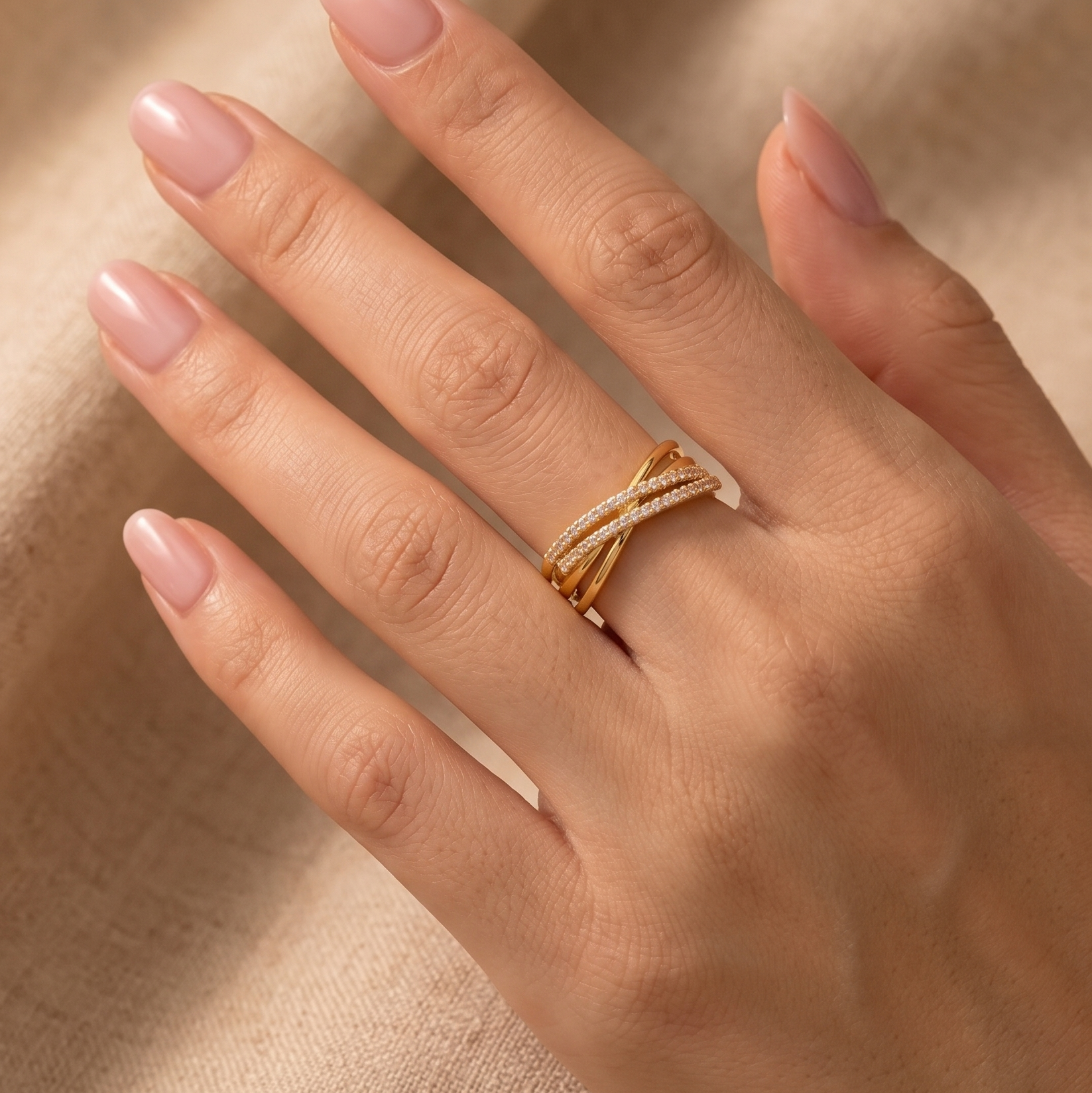 A close-up of a hand wearing a gold wrap ring with pavé-set clear stones, set against a soft beige textured fabric background.