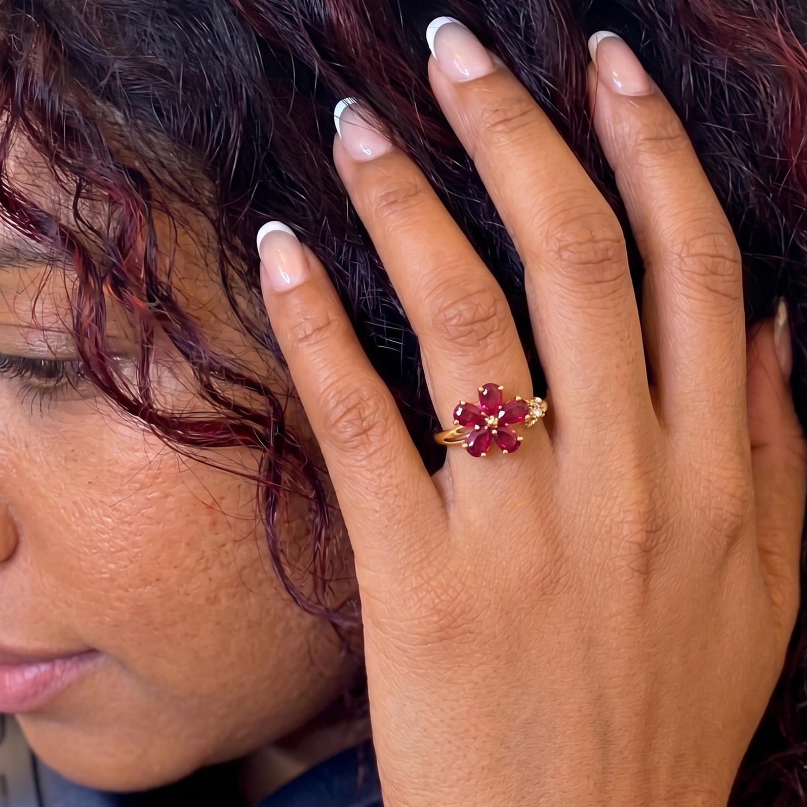 A close-up lifestyle shot of a person's hand wearing a gold ring with a flower-shaped cluster of deep red gemstones.