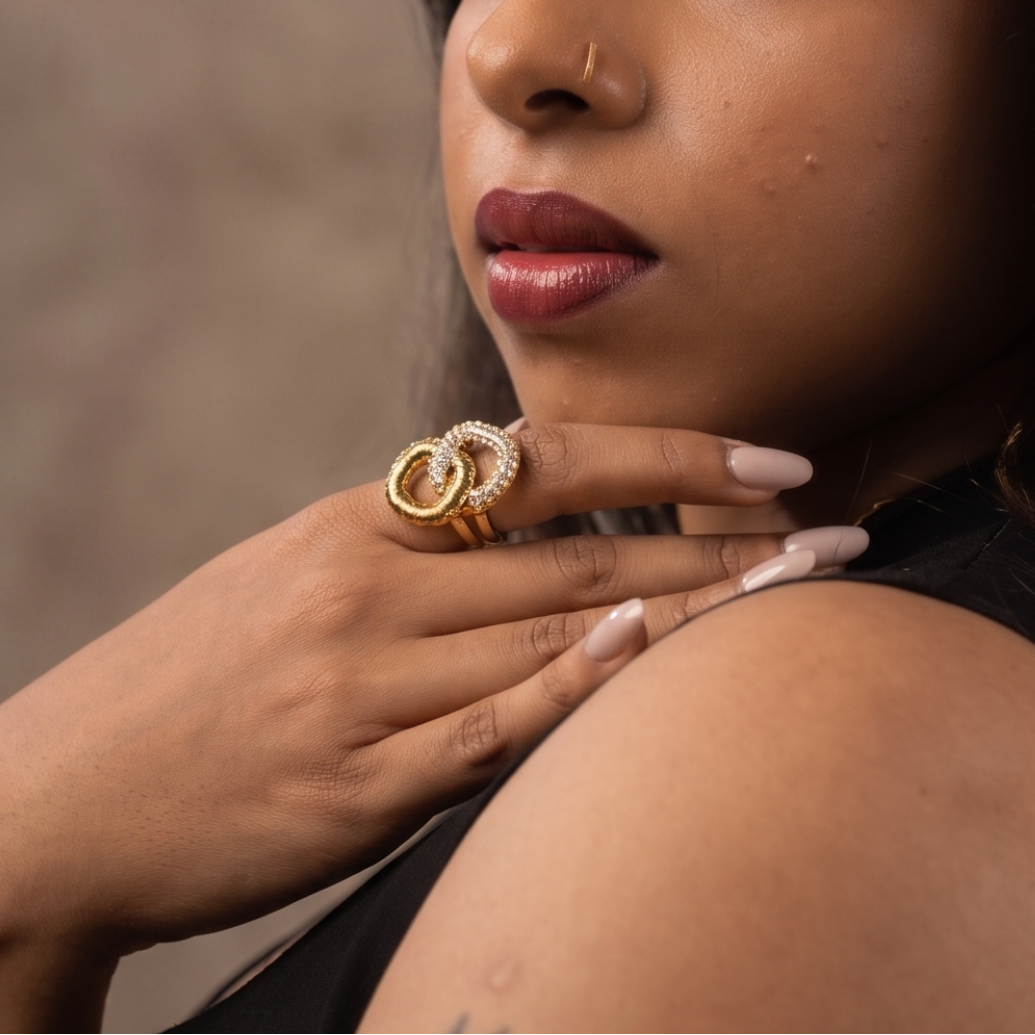 A woman with long dark hair is shown in a close-up portrait, wearing a large gold ring on her finger and a black sleeveless top. The background is a textured warm brown studio backdrop.
