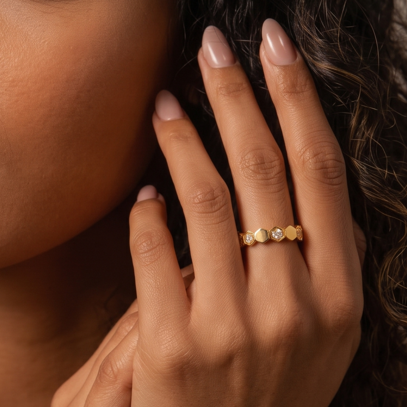A close-up lifestyle shot of a woman with dark curly hair wearing a delicate gold ring with small heart-shaped stones on her ring finger.